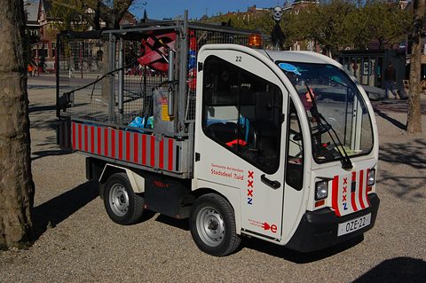 White narrow electric utility truck with red and white stripes and a cage body full of brooms on gravel under trees. Opens a larger view