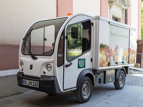 White narrow electric van with a box body printed with fruit and vegetables, parked beside a pink wall. Opens a larger view