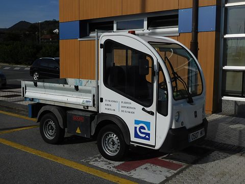 White narrow electric utility truck with Basque and Spanish lettering and a drop-side bed by a timber-clad building. Opens a larger view