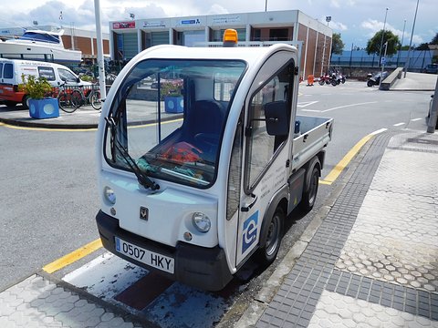 White narrow electric utility truck with an amber beacon and a drop-side bed on a quayside by a marina. Opens a larger view