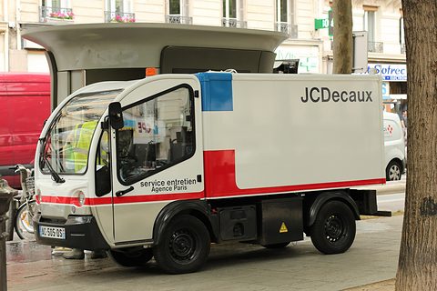 White and red narrow electric box van lettered JCDecaux parked under plane trees beside a street kiosk.