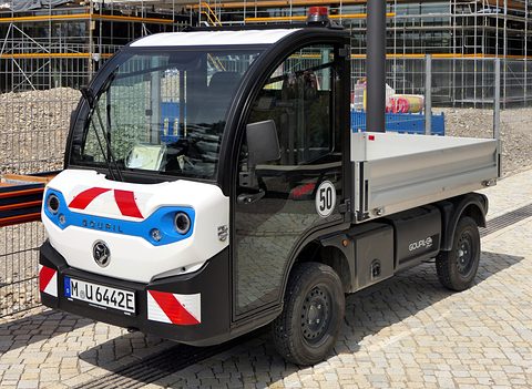 White and black narrow electric utility truck with a tipper bed and red and white chevrons on cobbles. Opens a larger view