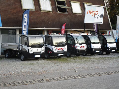 Five small black and white electric utility trucks lined up on gravel in front of a dealer’s building and flags. Opens a larger view