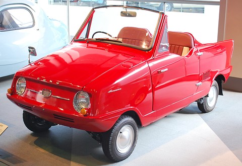 Small open red two-seater with a low windscreen, round headlamps and white wheels on a polished museum floor.