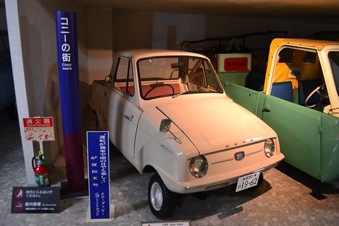 Cream pickup beside a green van in a dim museum bay, with a tall Cony town sign and display placards. Opens a larger view