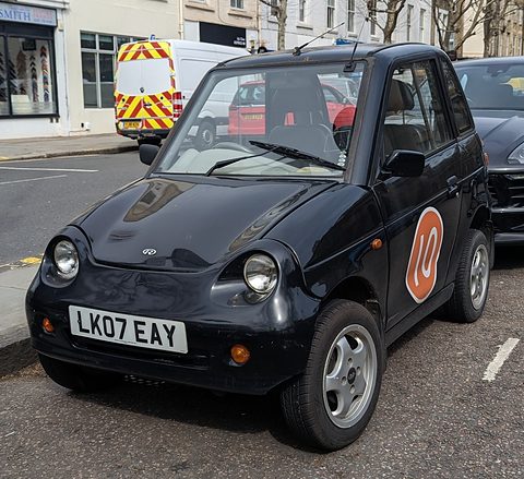 Front of a black tall microcar with round headlamps and an orange circular sticker on the door at a kerb. Opens a larger view