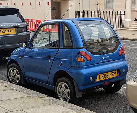 Rear of a blue tall microcar with round tail lamps and an LK56 plate, parked behind a Range Rover. Opens a larger view
