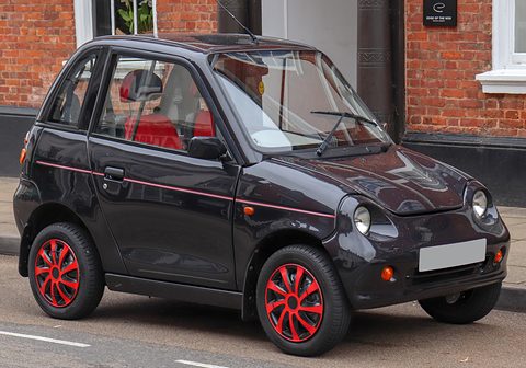 Black tall two-door microcar with red alloy wheels and a pinstripe, parked in front of a brick building. Opens a larger view