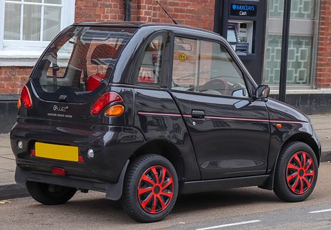 Rear of a black microcar with red wheels, round tail lamps and a yellow blanked plate beside a cash machine. Opens a larger view