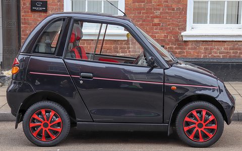Side view of a black, short and tall two-door microcar with red wheels and a red pinstripe by brick houses. Opens a larger view
