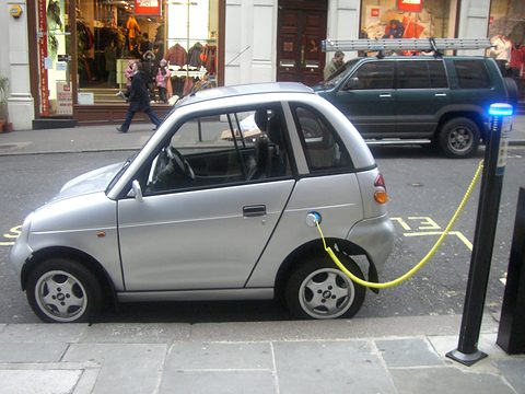 Silver microcar with a yellow charging cable to a black post with a blue light, shops and a 4x4 behind. Opens a larger view