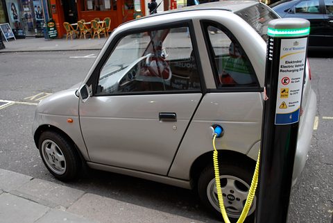 Silver microcar with a yellow cable running to a black Elektrobay charging post on a London pavement.