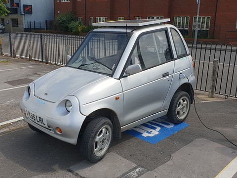 Silver tall two-door microcar with round headlamps parked over a blue painted charging-bay symbol by railings. Opens a larger view