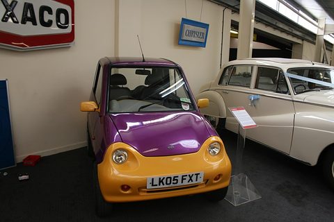 Purple microcar with a yellow front bumper and an LK05 plate beside a cream saloon and a Texaco sign. Opens a larger view