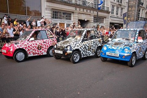 Three tall microcars in pink, zebra and blue graffiti liveries in a street lined with a crowd of spectators.