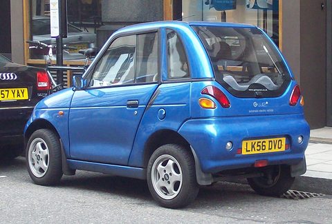 Rear three-quarter of a metallic blue microcar with round tail lamps and an LK56 plate at a city kerb. Opens a larger view