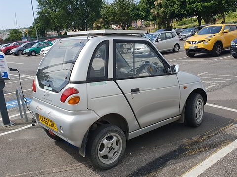 Rear of a short, tall silver two-door car with round tail lamps and a yellow UK plate in a car park. Opens a larger view