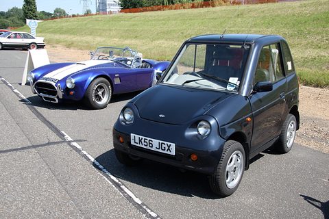 Dark blue tall microcar with an LK56 plate on tarmac, a blue and white-striped open sports car behind. Opens a larger view