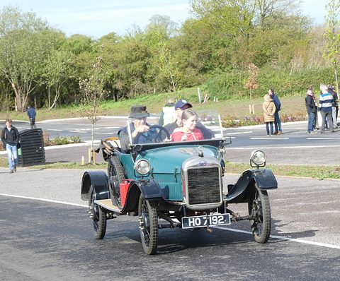 Front of a teal open two-seater with a flat radiator and two occupants, driving on a car park road. Opens a larger view