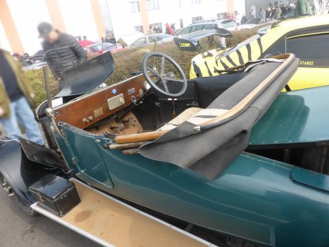 Open teal two-seater seen from the side, showing a wooden dashboard, steering wheel and folded hood. Opens a larger view