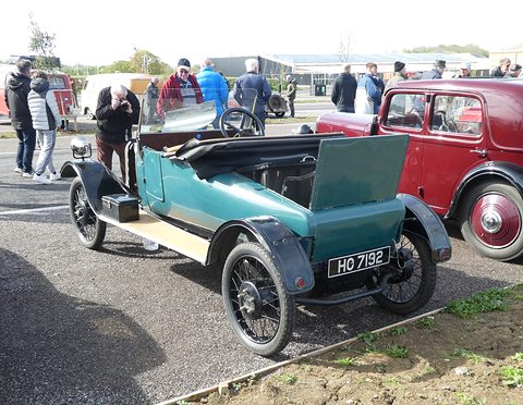 Teal two-seater with an open bonnet panel and black wings on gravel, a red saloon and visitors behind. Opens a larger view