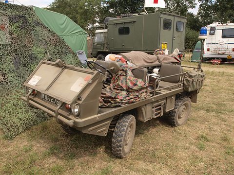 Olive small 4x4 carrying a stretcher and kit beside a camouflage net, an army ambulance behind. Opens a larger view