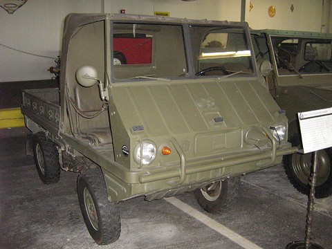 Olive military 4x4 with a soft top and a ribbed nose in a museum, another vehicle behind. Opens a larger view