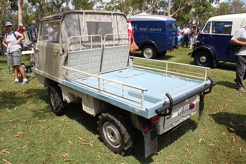 Rear of a grey small 4x4 with a pale blue load bed and rails, parked on grass among old vans. Opens a larger view