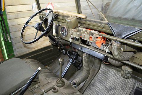 Steering wheel, gear lever and simple dashboard of a small military 4x4 seen from the open side. Opens a larger view
