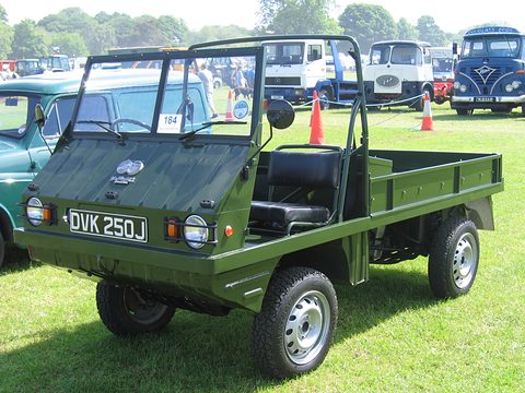 Green small open 4x4 with roll bars and a black seat on grass, lorries and buses behind. Opens a larger view