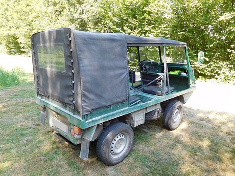 Rear of a green small 4x4 with a black canvas tilt standing on a meadow before trees. Opens a larger view