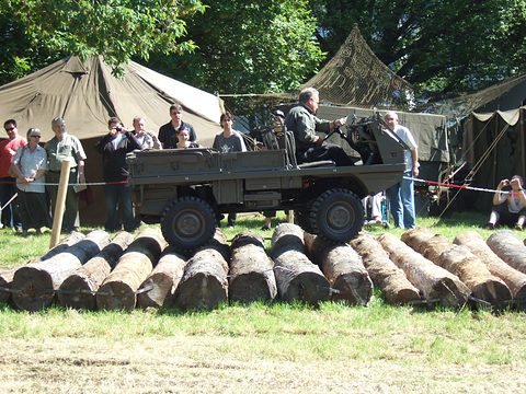 A small olive 4x4 with soldiers aboard crossing a row of logs, spectators and army tents behind. Opens a larger view