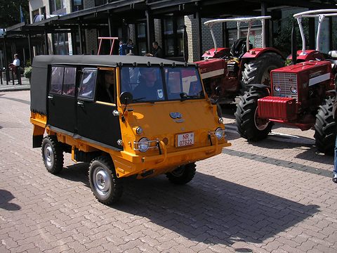 Orange small 4x4 with a black canvas crew cab parked on paving beside red tractors. Opens a larger view