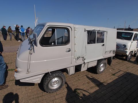 Grey small truck with a long rounded cab and a canvas load box, people standing behind. Opens a larger view