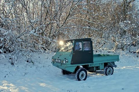 Green small truck with a rounded cab and lights on in deep snow among frosted trees. Opens a larger view
