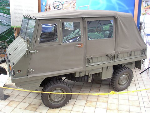 Olive small 4x4 with a full canvas cab behind a yellow rope on a tiled floor. Opens a larger view
