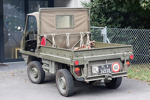 Rear of a small olive 4x4 with a canvas tilt and a Liechtenstein plate, parked by a fence. Opens a larger view