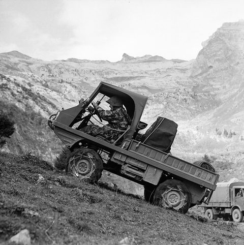 A small open military 4x4 with a soldier at the wheel tilted on a steep rocky hillside below mountains.