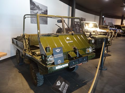 Olive small open 4x4 with a folded screen on a dark floor, a museum sign on its bumper. Opens a larger view