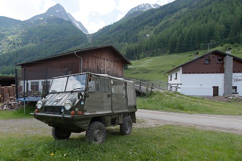 Olive small 4x4 with a canvas cab on a grass verge, a barn and snowy peaks behind.