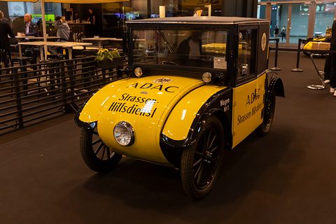A small yellow saloon with a black cab lettered ADAC Strassen-Hilfsdienst and black wire wheels on a dark show floor. Opens a larger view