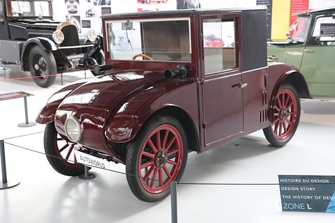 A small maroon saloon with a black cab roof, red wheels and an Autoworld sign before it, behind a rope. Opens a larger view