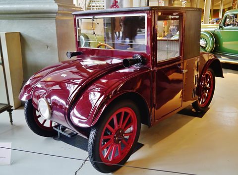A glossy maroon saloon with a bulb horn on the bonnet, a single headlamp and red wooden wheels. Opens a larger view