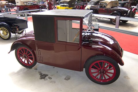 Side view of a small maroon saloon with a black fabric roof, one door and red wheels on a pale floor. Opens a larger view