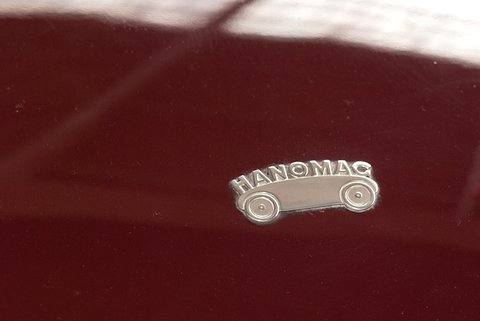 A small chrome badge shaped like a car with the word Hanomag, on glossy maroon paint. Opens a larger view
