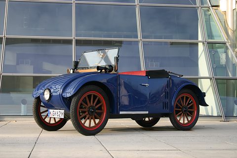 A dark blue open two-seater with red wooden wheels and a single headlamp before a curved glass building. Opens a larger view