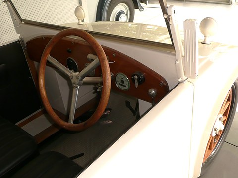 A wooden steering wheel, a mahogany dashboard with a few gauges and a black bench seat inside a cream car. Opens a larger view
