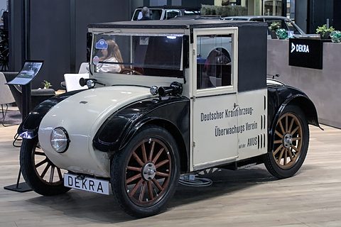 A tiny black and cream van with a lettered box body, wooden wheels and a single round headlamp on a show stand. Opens a larger view