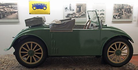 Side view of a green two-seater with cream spoked wheels and a suitcase on its tail, before framed factory photographs. Opens a larger view