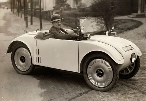 A man in a hat sits in a small pale open two-seater with a single round headlamp in its rounded nose.